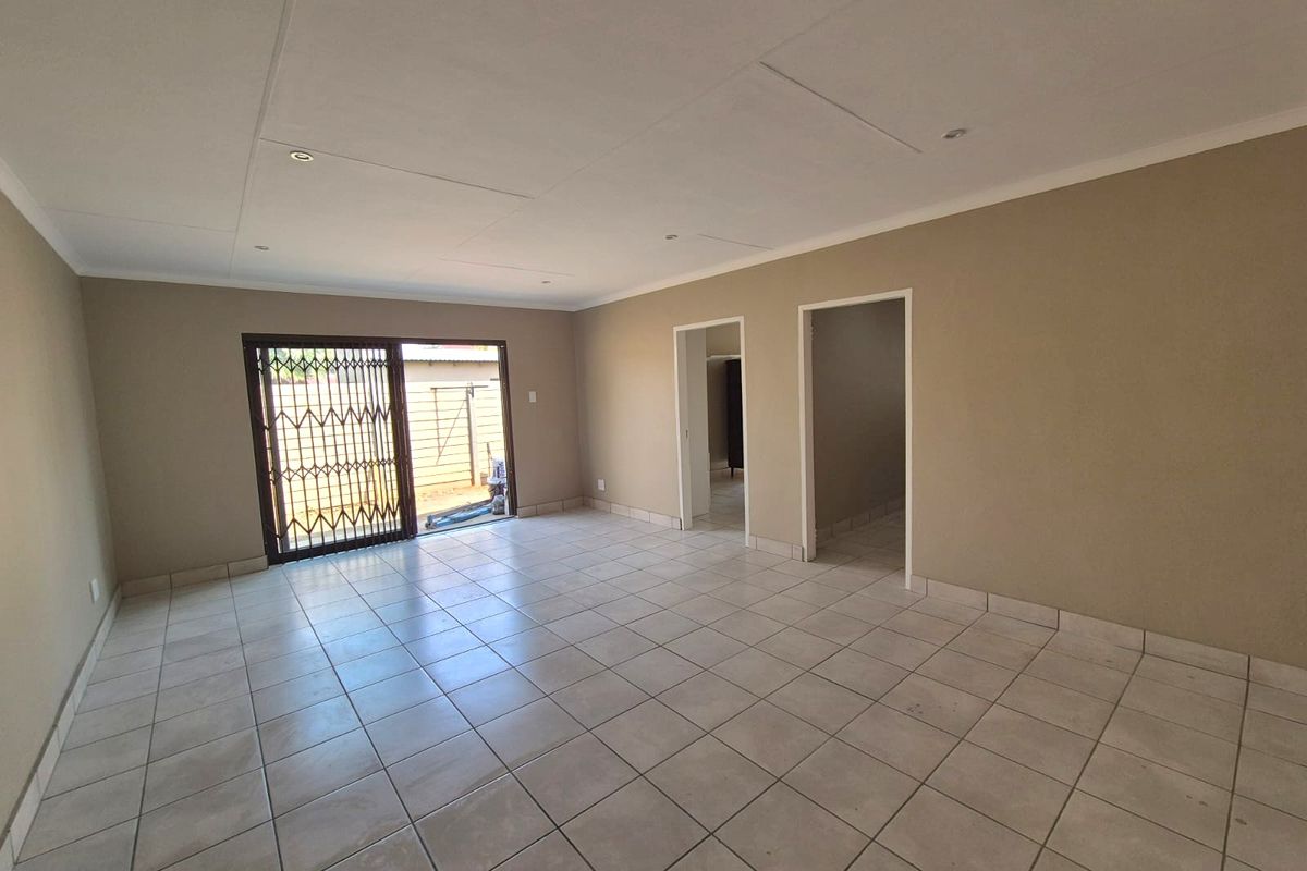 A spacious, empty living area featuring neutral tiled flooring, recessed lighting, and a sliding glass door with a security gate leading outdoors.