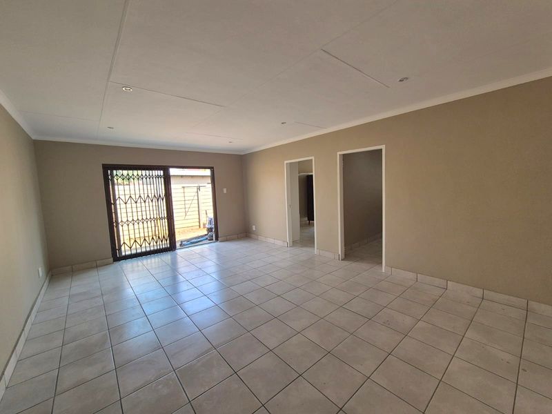 A spacious, empty living area featuring neutral tiled flooring, recessed lighting, and a sliding glass door with a security gate leading outdoors.
