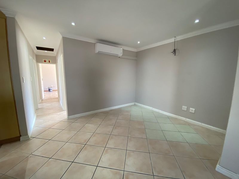 An empty living area featuring ceramic tiled flooring, recessed lighting, and a wall-mounted air conditioning unit, with an adjacent hallway visible.