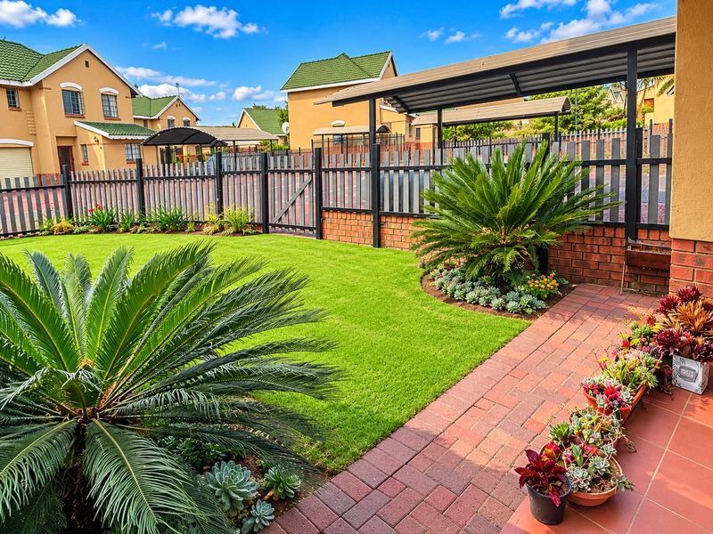 A well-maintained rear garden featuring brick paving, a covered patio area, and secure metal perimeter fencing within a townhouse complex.