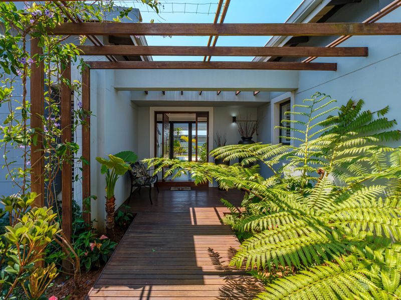 Inviting entrance featuring a timber deck walkway, overhead pergola structure, and a modern glass-panelled front door surrounded by lush garden landscaping.