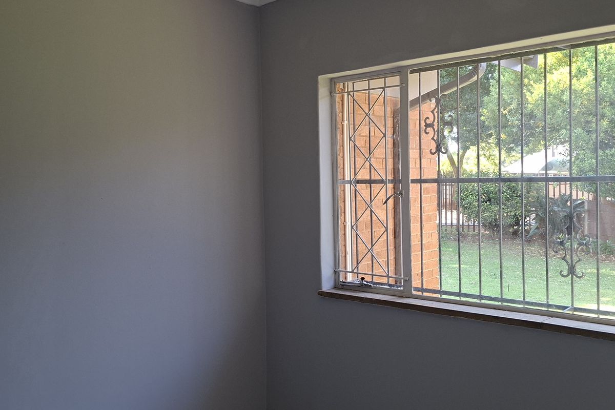 A bright, empty bedroom featuring neutral walls and a large window fitted with security bars, overlooking the garden.