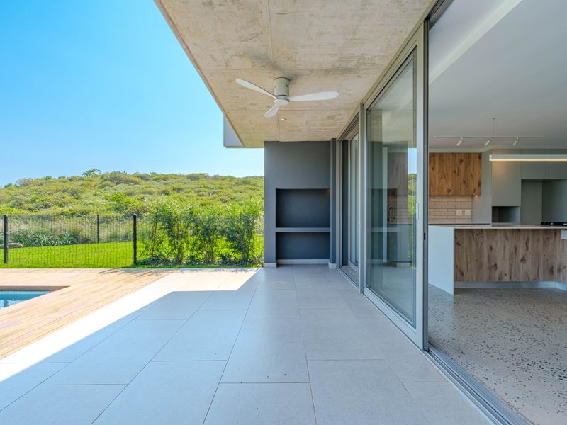 Modern covered patio featuring an exposed concrete ceiling, built-in braai, and seamless flow to the swimming pool and timber deck area.