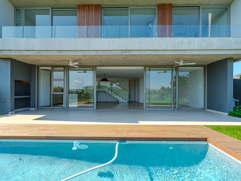 Modern outdoor entertainment area featuring a swimming pool with timber decking, a spacious tiled patio, built-in braai, and seamless indoor-outdoor flow through large sliding doors.
