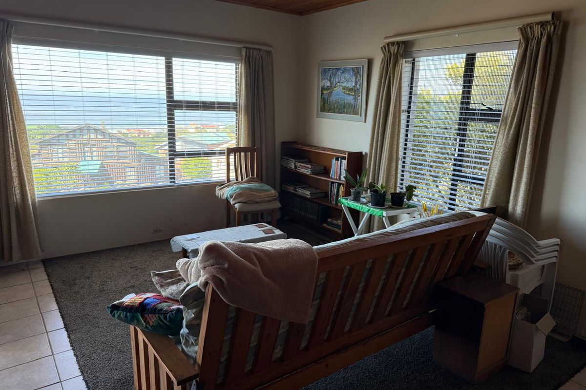 A living room featuring a timber ceiling, large windows with venetian blinds, and a combination of tiled and carpeted flooring with coastal views.