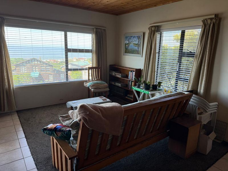 A living room featuring a timber ceiling, large windows with venetian blinds, and a combination of tiled and carpeted flooring with coastal views.