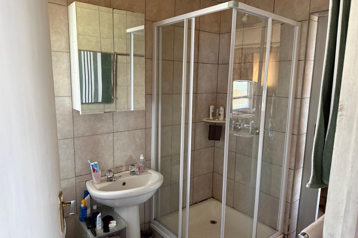 A functional bathroom featuring a corner shower enclosure, pedestal washbasin, and a wall-mounted mirrored medicine cabinet with neutral ceramic wall tiles.