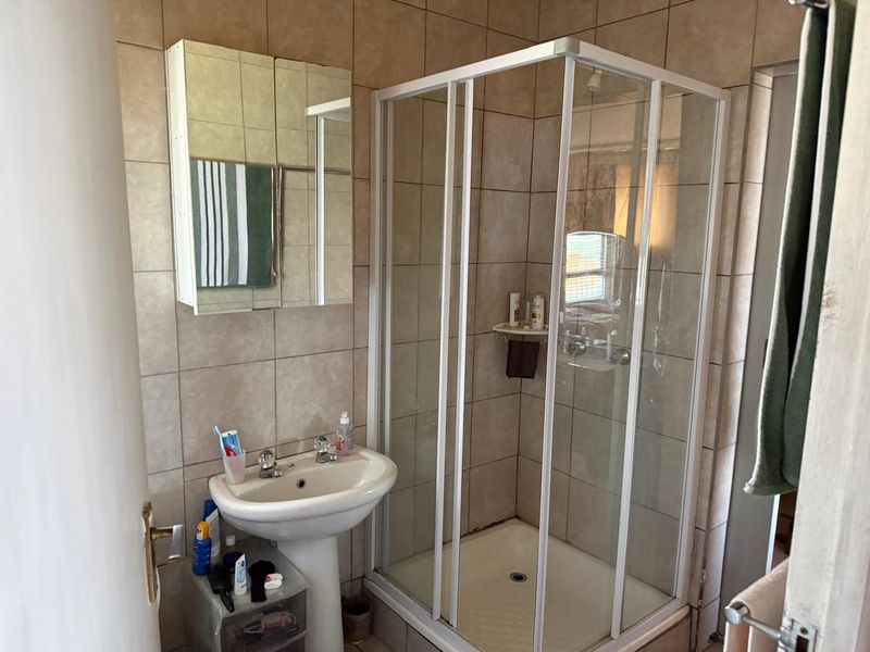 A functional bathroom featuring a corner shower enclosure, pedestal washbasin, and a wall-mounted mirrored medicine cabinet with neutral ceramic wall tiles.