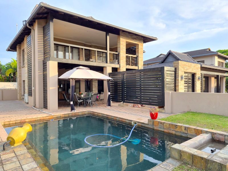 A well-maintained outdoor pool area featuring a paved deck, stone-clad accents, a timber privacy screen, and a covered patio with an upper-level balcony.