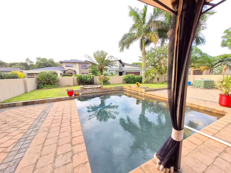 A well-maintained outdoor pool area featuring a paved patio, surrounding boundary walls, and a covered gazebo structure overlooking the garden.