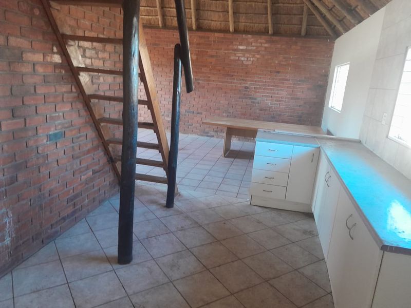 Open plan living area featuring exposed brick walls, a traditional thatch roof, tiled flooring, and a basic kitchen setup with a wooden staircase.