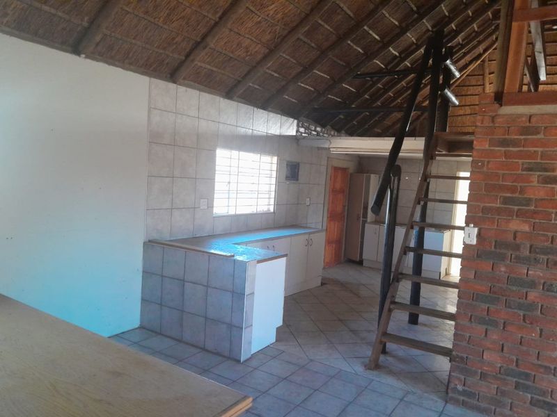 Open-plan kitchen area featuring tiled counters, exposed brickwork, and a wooden staircase leading to a mezzanine level under a traditional thatch roof.