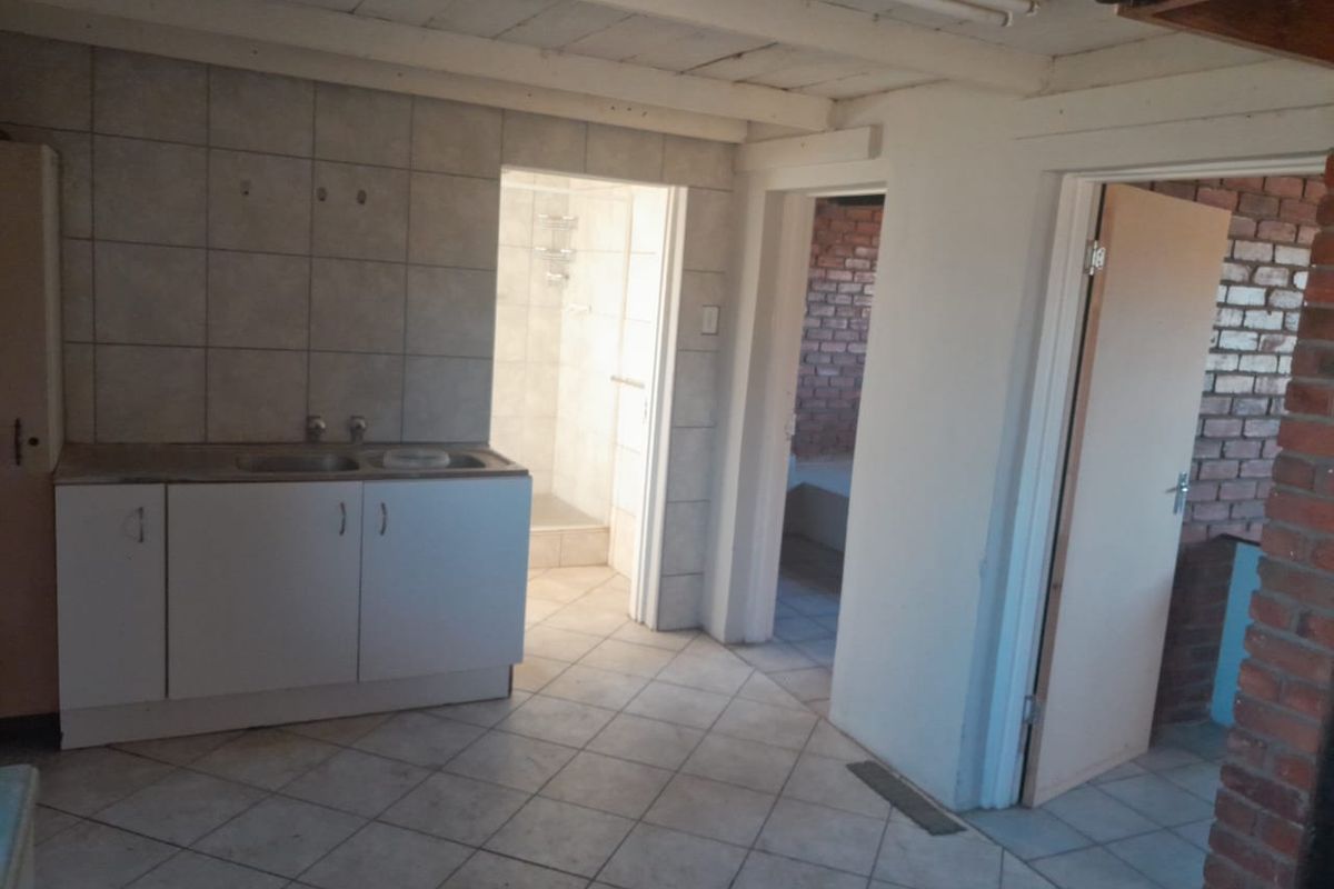 A basic kitchen area featuring tiled flooring, cabinetry with a double sink, and exposed timber ceiling beams, with direct access to an adjacent bathroom.