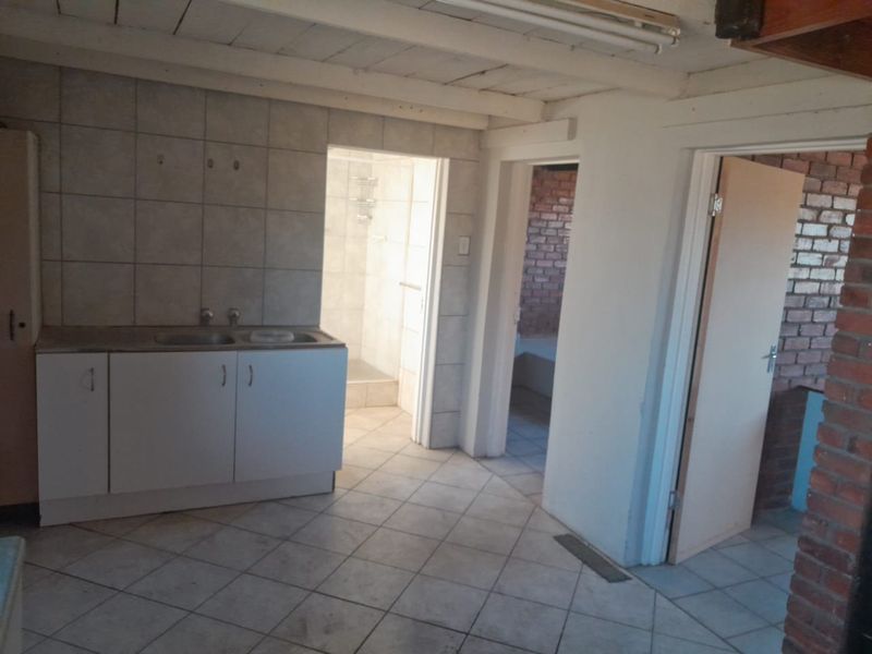 A basic kitchen area featuring tiled flooring, cabinetry with a double sink, and exposed timber ceiling beams, with direct access to an adjacent bathroom.