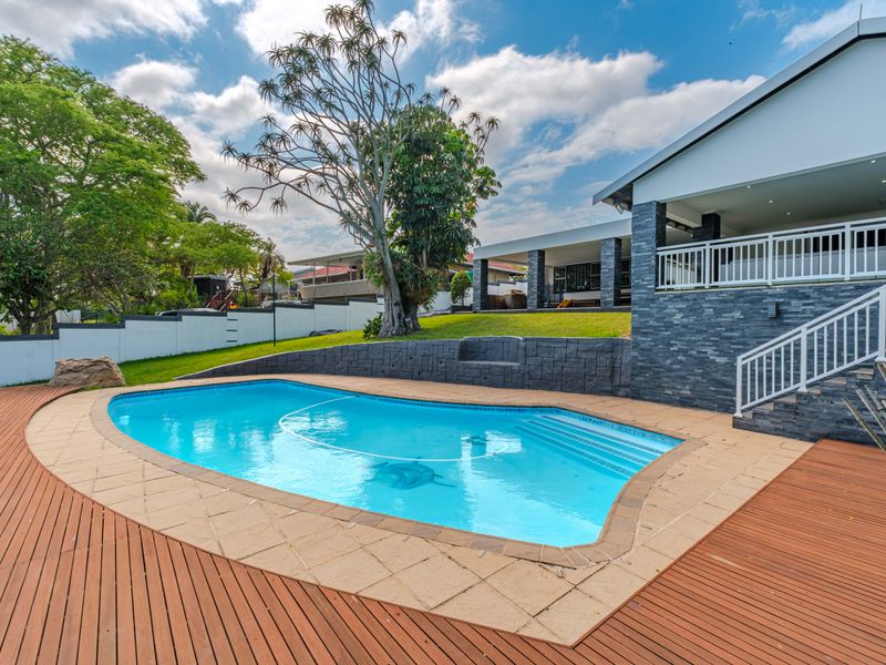 A well-maintained outdoor pool area featuring timber decking, stone-clad retaining walls, and a staircase leading to a covered patio.