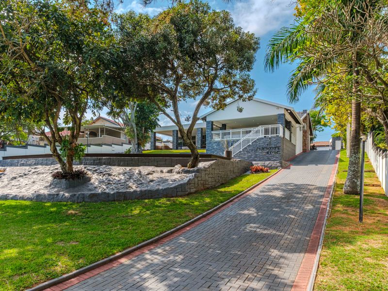 Front view of the property featuring a paved driveway, landscaped garden with retaining walls, and a covered entrance patio.