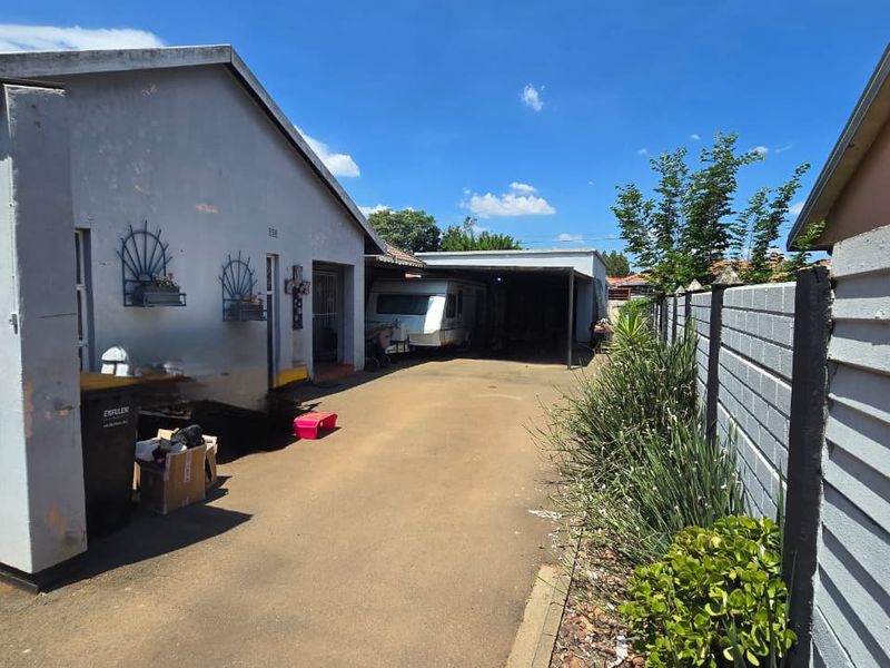 A rear exterior view showing a concrete driveway leading to a covered carport area, flanked by a boundary wall and garden border.