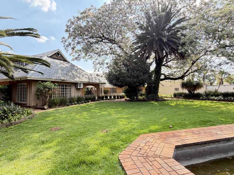 Spacious rear garden featuring a large lawn, mature trees, and a swimming pool with brick coping, adjacent to the main house structure.