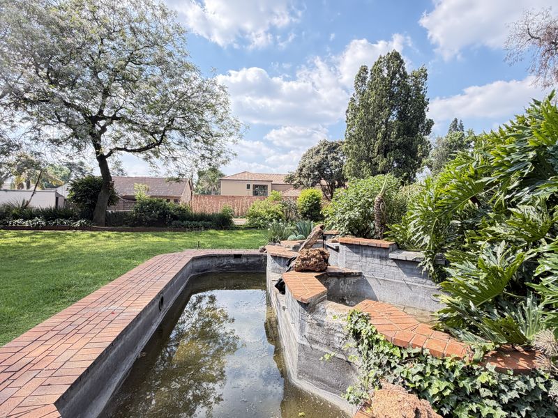 A spacious garden featuring a brick-edged water feature, mature trees, and a well-maintained lawn, providing a tranquil outdoor setting.