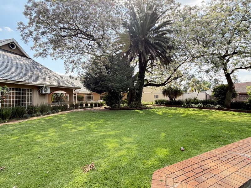 A spacious rear garden featuring a well-maintained lawn, mature trees, and a brick-edged swimming pool, adjacent to the main house structure.