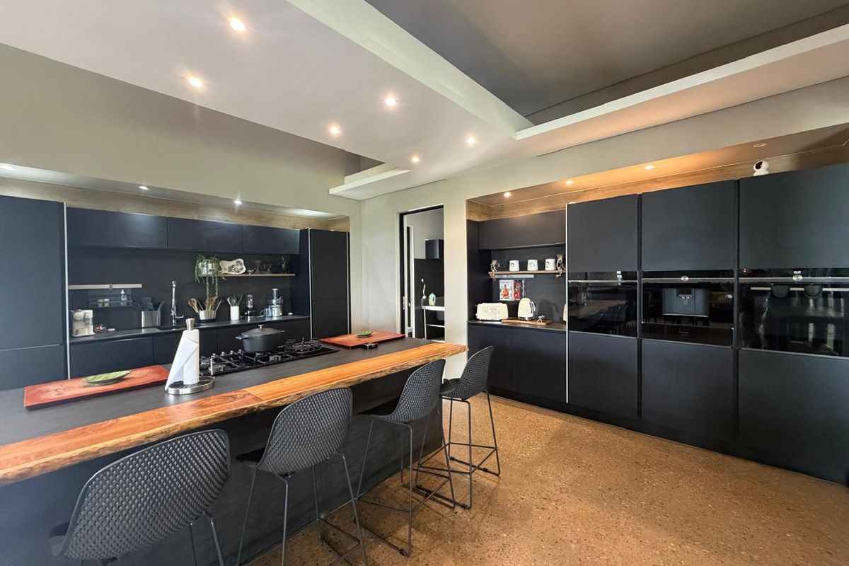 Modern kitchen featuring sleek black cabinetry, a large island with a live-edge wood breakfast bar, integrated appliances, and polished terrazzo flooring.