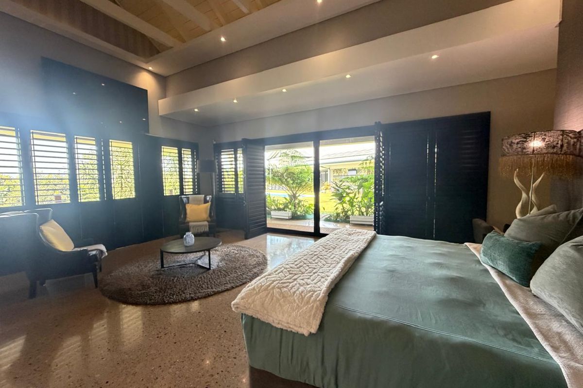 Spacious bedroom featuring vaulted ceilings with exposed beams, polished terrazzo flooring, and black plantation shutters opening to a private garden view.