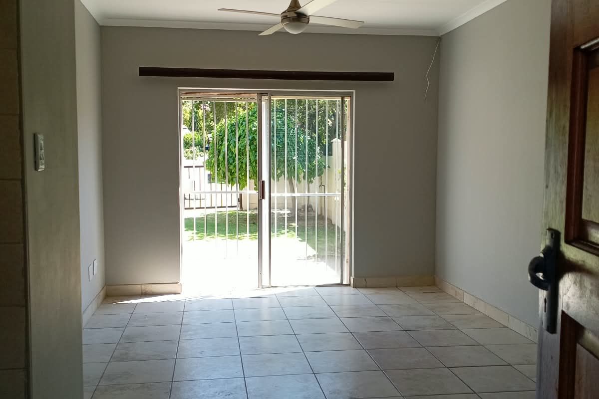 A bright, empty living room featuring neutral tiled flooring, a ceiling fan, and a sliding glass door providing direct access to the garden.