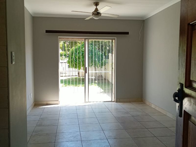 A bright, empty living room featuring neutral tiled flooring, a ceiling fan, and a sliding glass door providing direct access to the garden.