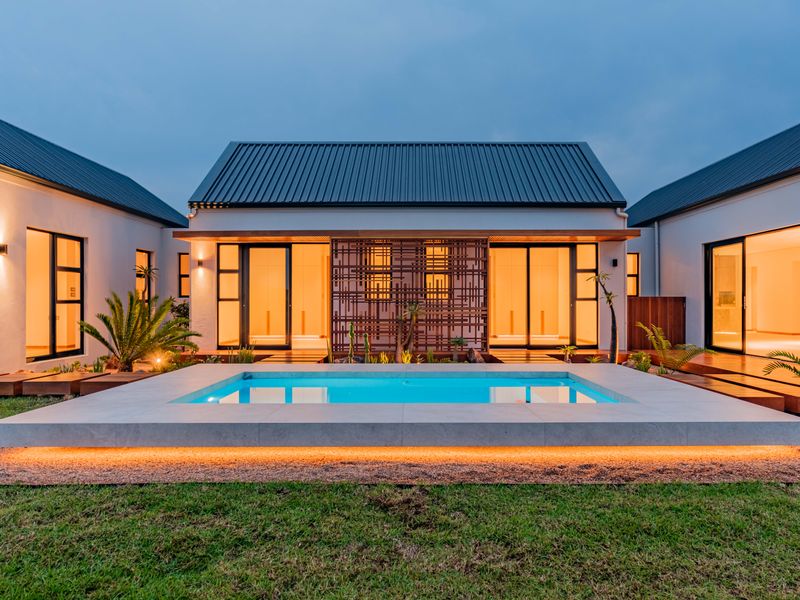 A modern architectural home featuring a pristine swimming pool, timber decking, and illuminated sliding glass doors, set within a beautifully landscaped garden.