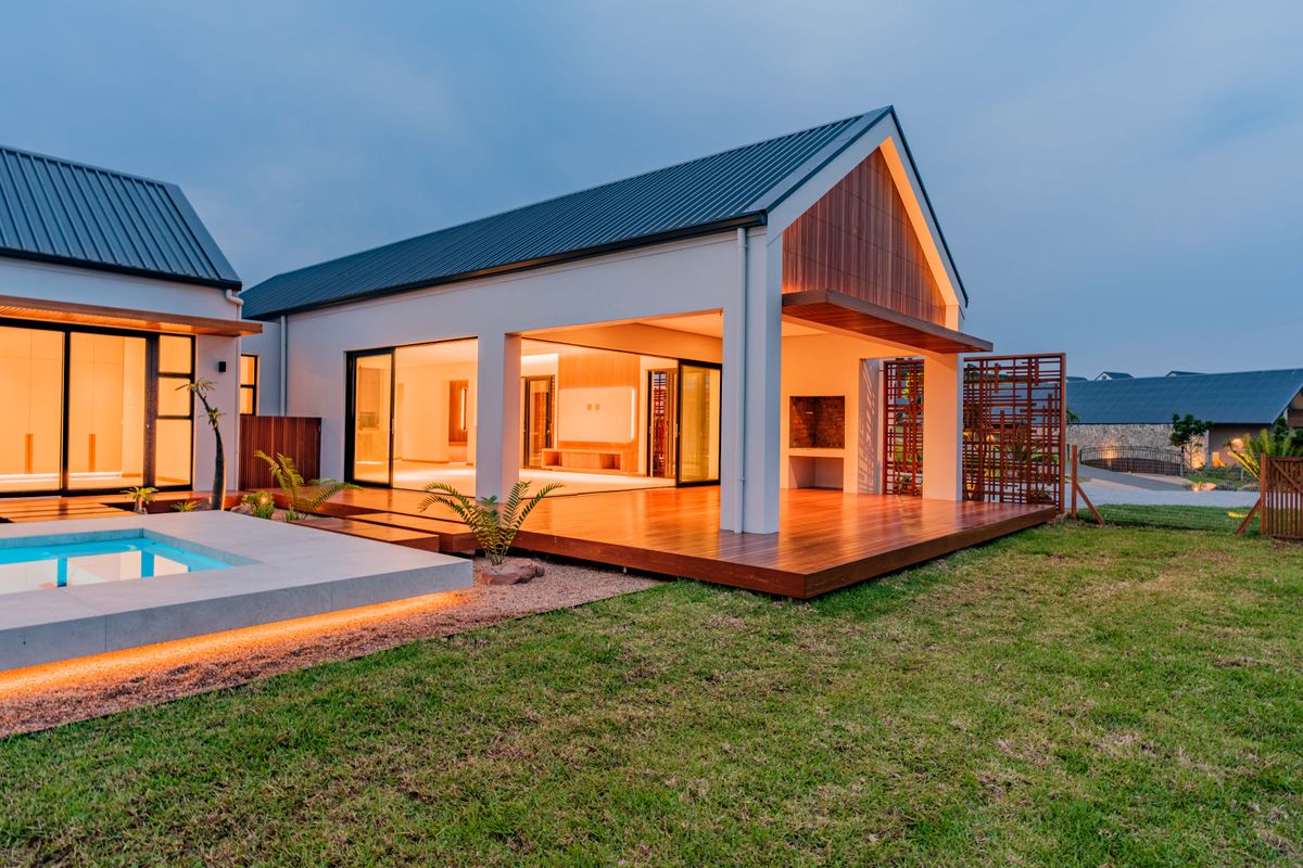 Modern rear exterior featuring a swimming pool, expansive timber decking, built-in braai area, and seamless indoor-outdoor flow through large sliding glass doors.
