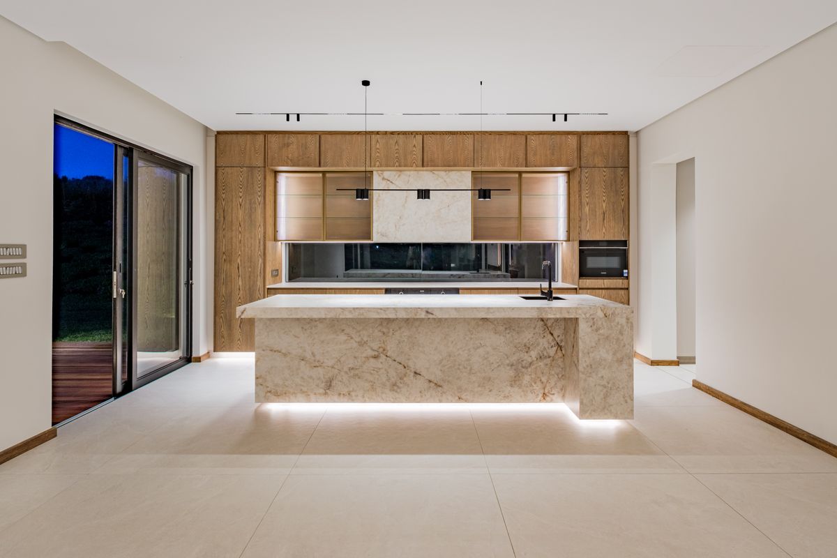 Modern luxury kitchen featuring a large stone island, integrated wood cabinetry, built-in appliances, and sleek recessed lighting with direct access to an outdoor area.