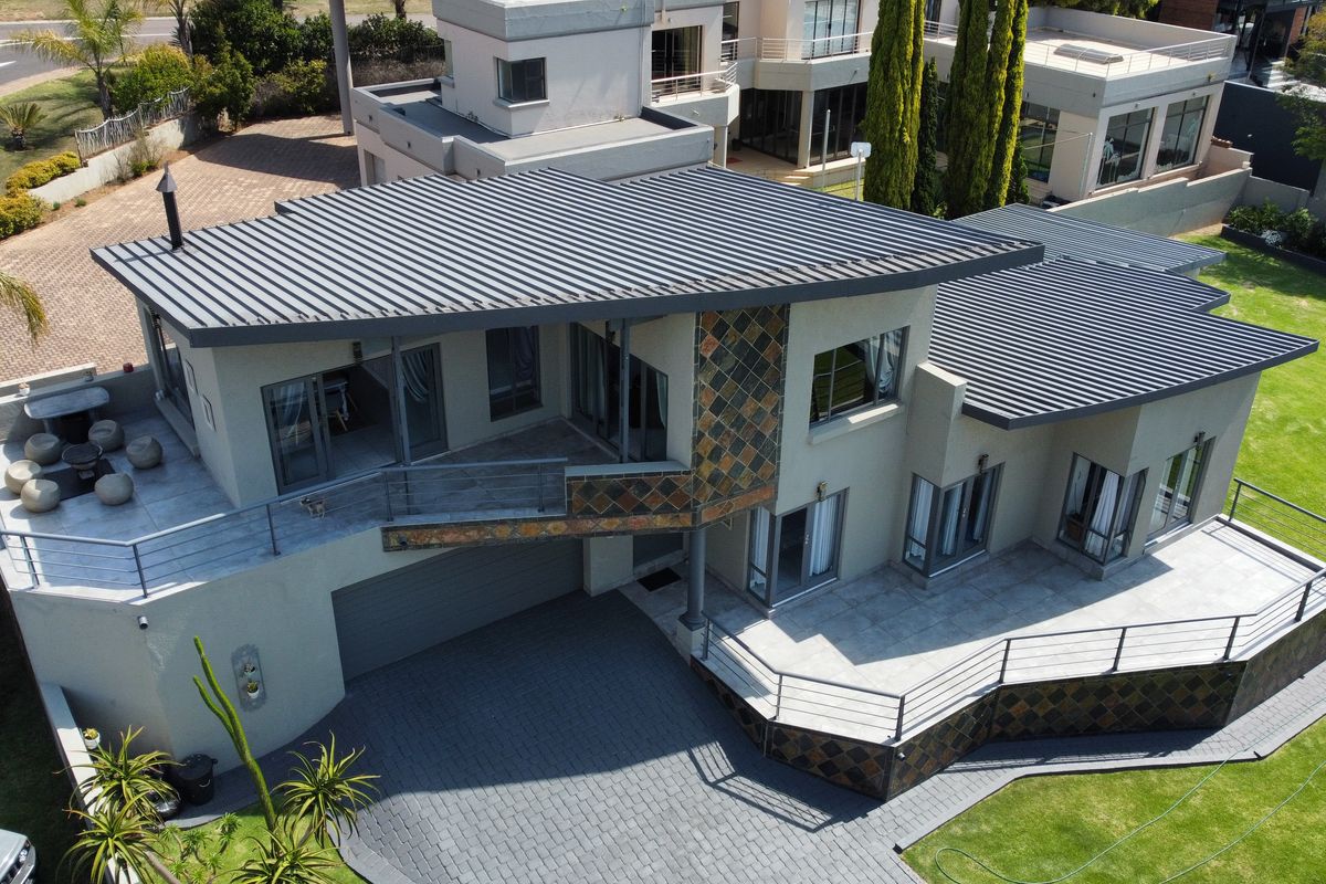 Aerial view of a modern multi-level residence featuring a metal roof, stone-clad accents, paved driveway, and spacious balconies overlooking a well-maintained garden.