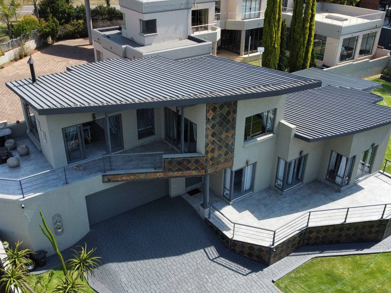 Aerial view of a modern multi-level residence featuring a metal roof, stone-clad accents, paved driveway, and spacious balconies overlooking a well-maintained garden.