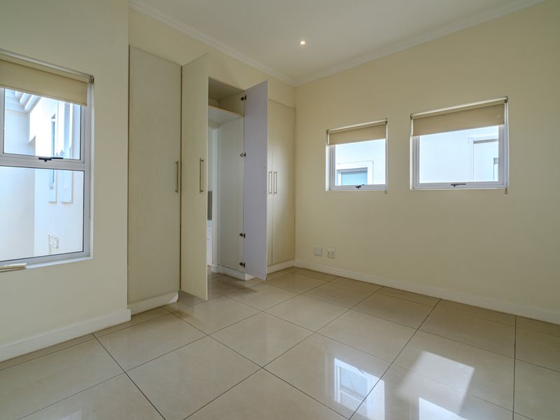 A bright, empty bedroom featuring neutral tiled flooring, built-in cupboards, and multiple windows with roller blinds, offering a clean and versatile living space.