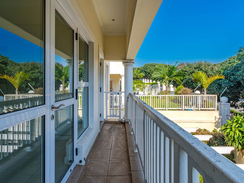 A covered balcony featuring tiled flooring, a white metal balustrade, and sliding glass door access, overlooking a landscaped garden area.