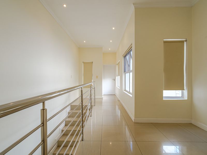A bright and spacious upper-level hallway featuring tiled flooring, a modern stainless steel balustrade, and neutral wall finishes with recessed lighting.