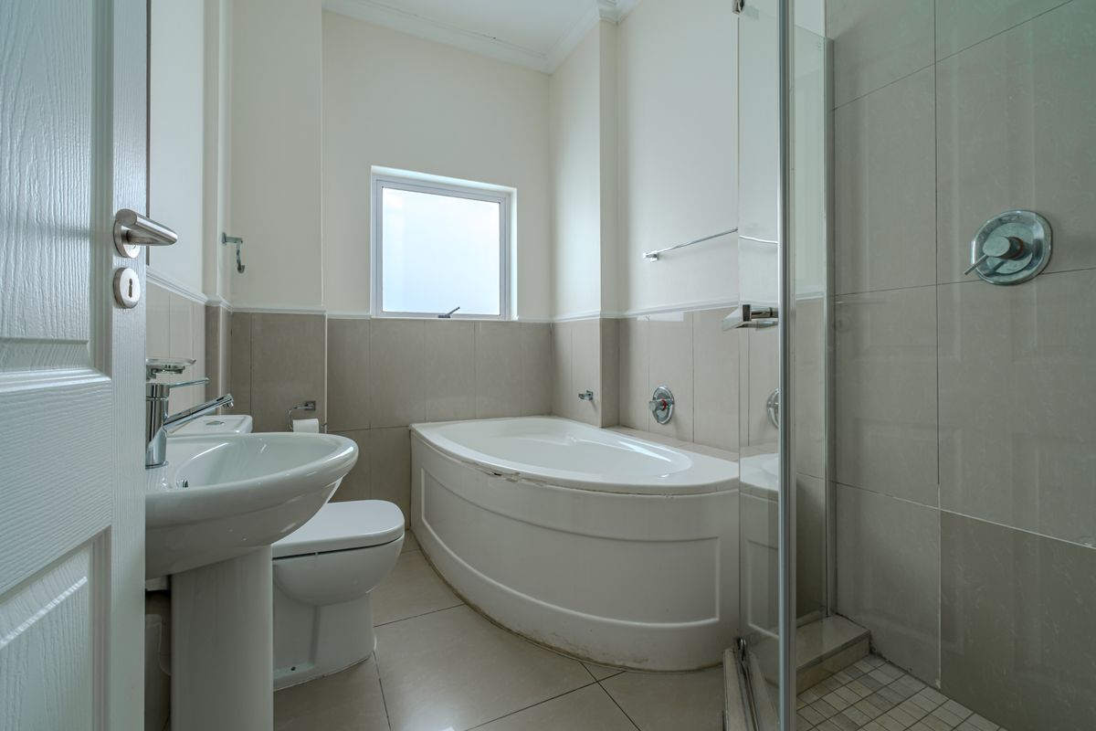 A clean, functional bathroom featuring a pedestal basin, toilet, curved bathtub, and a separate glass-enclosed shower with neutral wall and floor tiling.