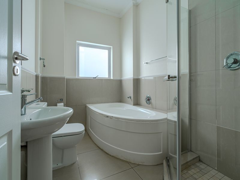 A clean, functional bathroom featuring a pedestal basin, toilet, curved bathtub, and a separate glass-enclosed shower with neutral wall and floor tiling.