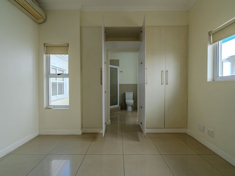 A bright, empty bedroom featuring tiled flooring, built-in cupboards, an air conditioning unit, and direct access to an en-suite bathroom.