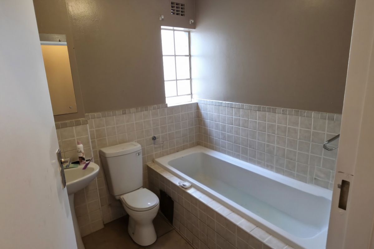 A compact bathroom featuring a bathtub, pedestal basin, and toilet with neutral wall tiles and a security-grille window.
