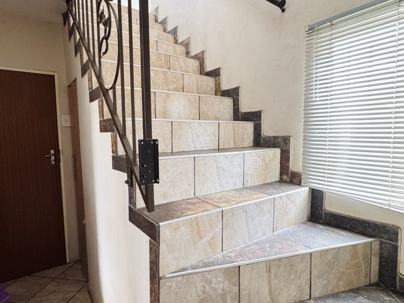 A functional hallway featuring a tiled staircase with a decorative wrought iron balustrade and natural light provided by a window with venetian blinds.