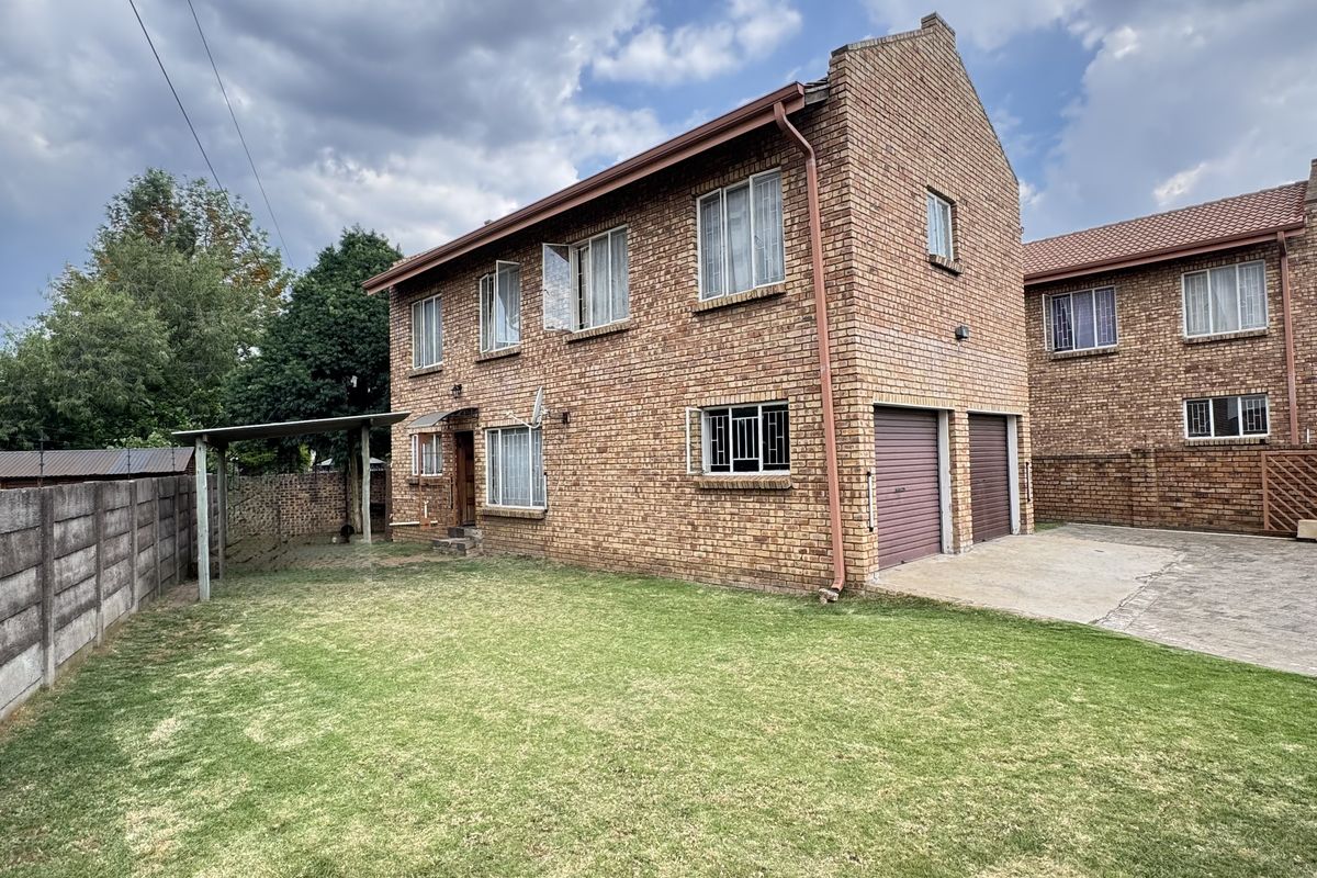 Rear view of a double-story face brick house featuring a double garage, concrete driveway, and a spacious lawn with a covered patio area.