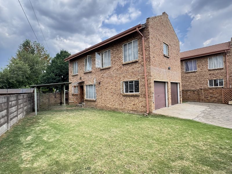 Rear view of a double-story face brick house featuring a double garage, concrete driveway, and a spacious lawn with a covered patio area.