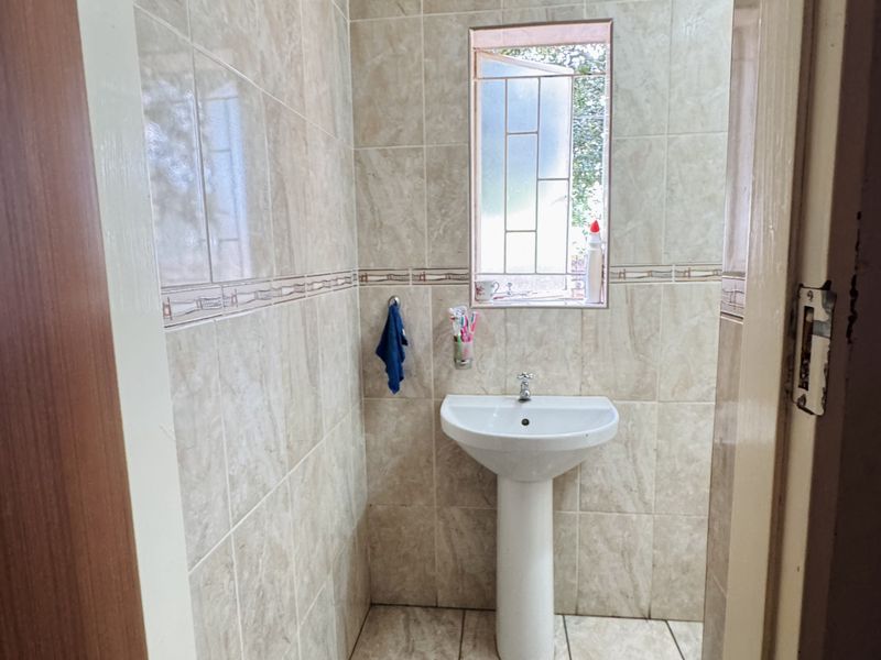 A compact bathroom featuring a pedestal basin, neutral ceramic wall and floor tiling, and a functional casement window for natural light.