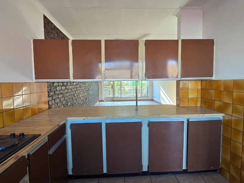 A dated kitchen featuring brown cabinetry, ceramic wall tiles, and an exposed brick wall, opening into a bright living area.
