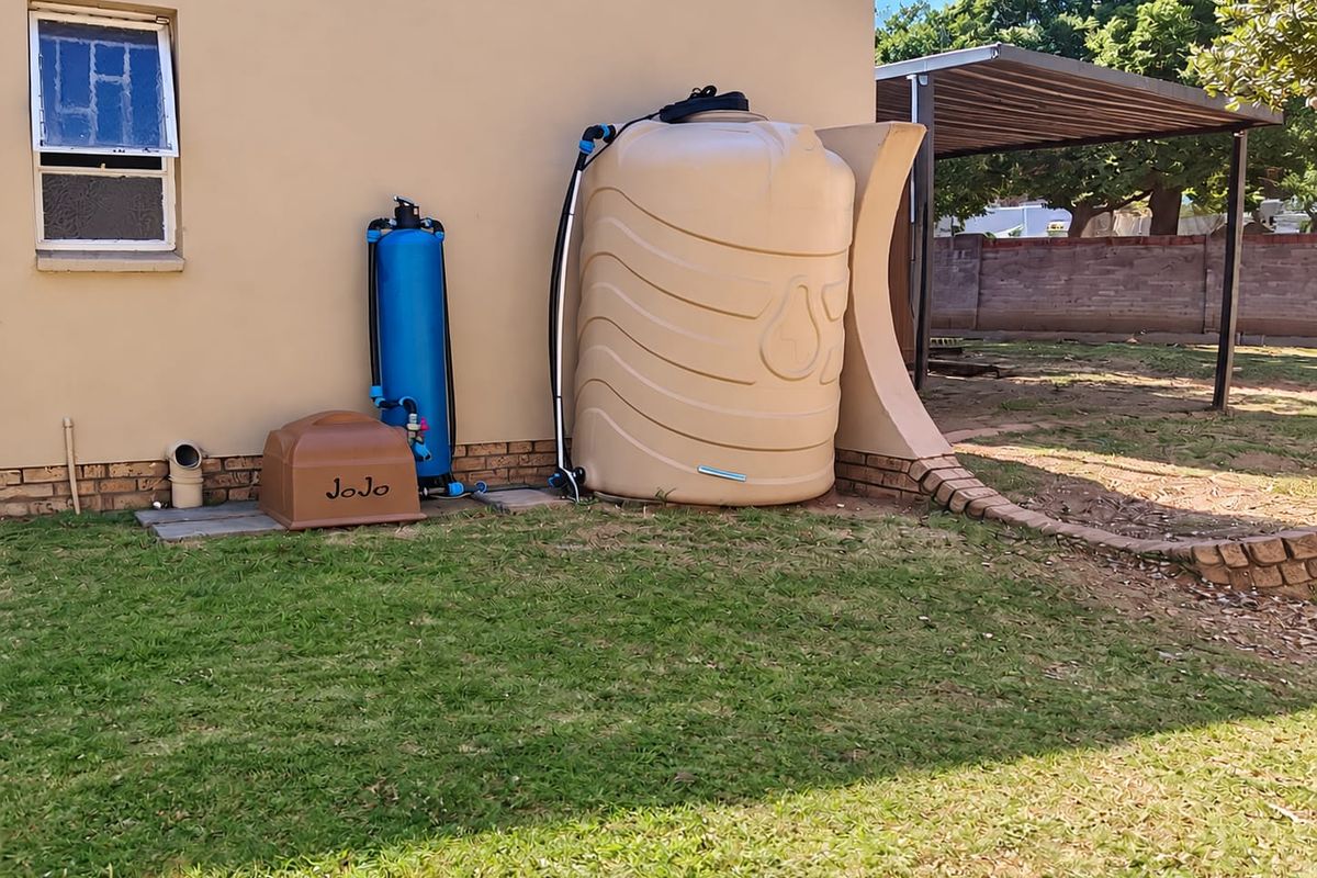 Rear exterior view featuring a water storage tank, filtration system, and a carport structure set within a landscaped garden area.