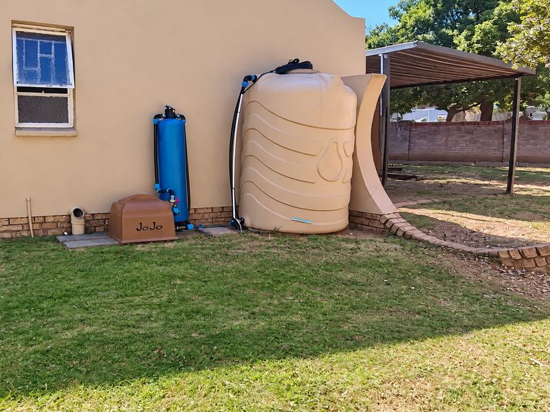 Rear exterior view featuring a water storage tank, filtration system, and a carport structure set within a landscaped garden area.