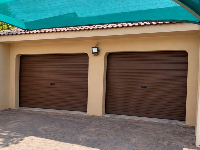A well-maintained double garage featuring brown roller doors, a paved driveway, and a protective shade sail structure.