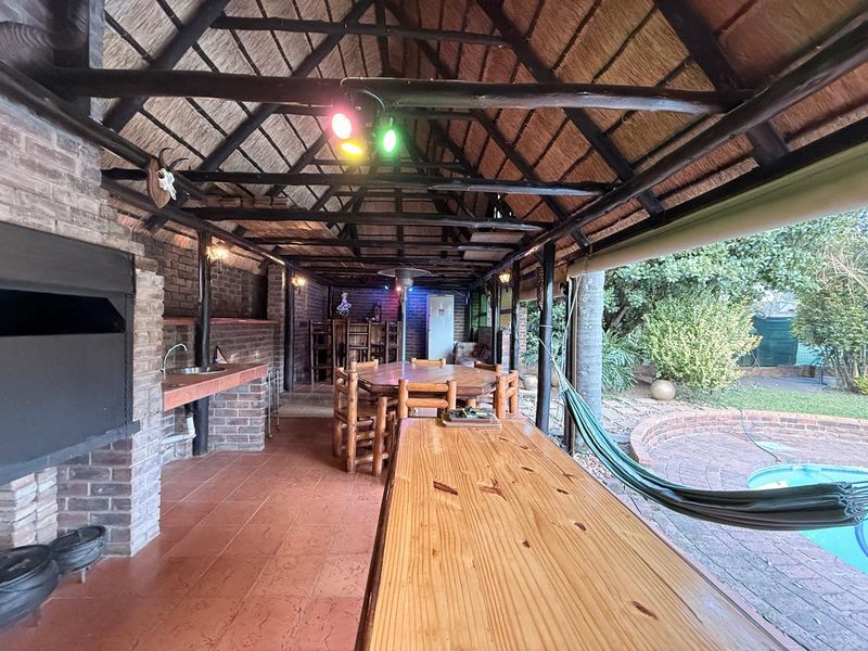 Spacious covered patio featuring a traditional thatched roof, exposed timber beams, built-in brick braai, and a prep area overlooking the garden and pool.