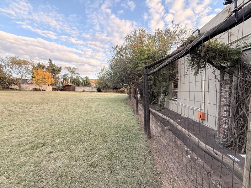 A spacious backyard featuring a large lawn area, perimeter wire fencing, and a paved walkway alongside the exterior wall of the house.